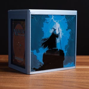 May include: A silver-framed card storage box with a blue illustration of a wizard holding a staff, silhouetted against a stormy sky. The box has a card visible inside with the word "MAGIC" on it. The box is on a wooden surface.
