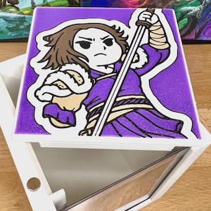 May include: A white box with a hinged lid featuring a cartoon illustration of a character in purple clothing holding a sword. The character has brown hair and is set against a textured purple background. The box is open, revealing a mirrored interior.