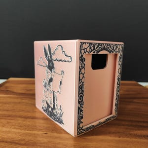 May include: A pink box with a black and white illustration of a rabbit holding a scroll. The box has a handle cutout and a decorative black border. The box is sitting on a wooden surface.