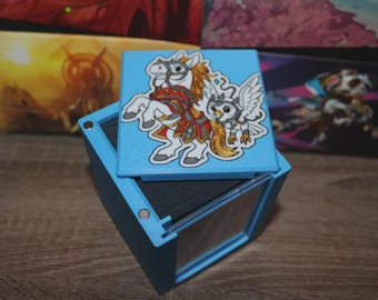 May include: A light blue box with a hinged lid, featuring a cartoon illustration of a white horse and a winged owl. The box is open, revealing a dark interior. Other boxes with colorful designs are visible in the background.