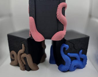 May include: Three black square boxes with textured surfaces, each adorned with colorful tentacles. One box features pink tentacles, another has brown tentacles, and the third displays blue tentacles. The boxes are arranged in a group.