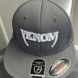 May include: A gray baseball cap with a white "VENOM" logo embroidered on the front. The cap has a black Flexfit label with the text "THE ORIGINAL FLEXFIT L/XL IT FITS YOUR LIFE" and a black CapAmerica label with the text "CA PREMIUM LINE".