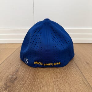 May include: A royal blue baseball cap with a curved peak. The cap has small ventilation holes and the words "GRIZZLY SPORT GEAR" in yellow. A white logo is on the side.