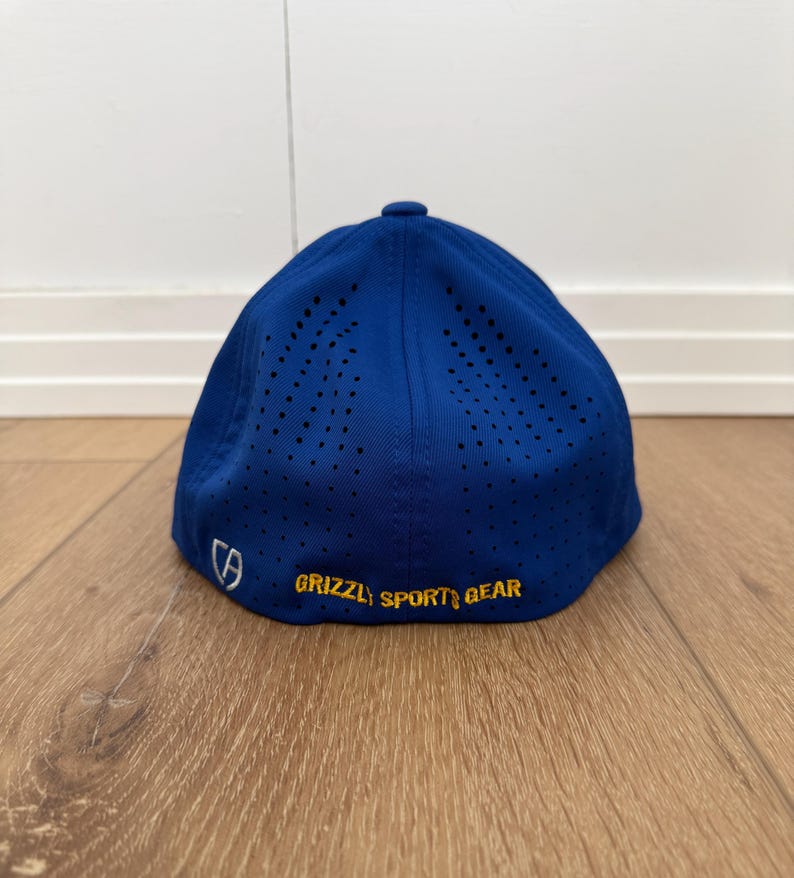 May include: A royal blue baseball cap with a curved peak. The cap has small ventilation holes and the words "GRIZZLY SPORT GEAR" in yellow. A white logo is on the side.