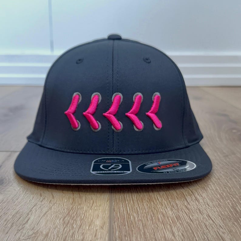 May include: A navy blue baseball cap with a curved peak. The cap has a series of pink embroidered chevrons across the front. The cap has a Flexfit sticker on the peak.
