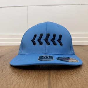 May include: A light blue baseball cap with a curved peak. The cap has a series of dark blue chevron designs embroidered on the front. The cap is on a wooden surface.