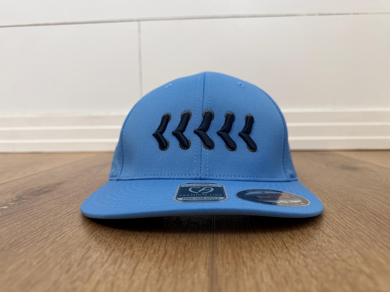 May include: A light blue baseball cap with a curved peak. The cap has a series of dark blue chevron designs embroidered on the front. The cap is on a wooden surface.