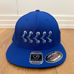 May include: A royal blue baseball cap with white embroidered design. The cap features a flat brim and a structured crown. The front of the cap has a white embroidered design. The cap has a Flexfit L/XL label.