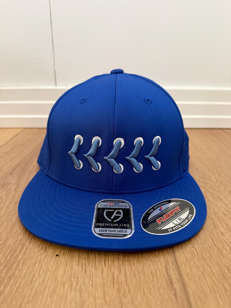 May include: A royal blue baseball cap with white embroidered design. The cap features a flat brim and a structured crown. The front of the cap has a white embroidered design. The cap has a Flexfit L/XL label.