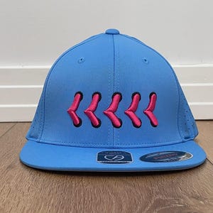 May include: A sky blue baseball cap with a flat peak. The cap has a series of pink embroidered chevrons across the front. The cap has a structured fit and a small logo on the brim.