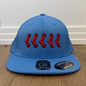 May include: A light blue baseball cap with a curved peak. The cap has six red, stylized chevrons embroidered across the front. The cap has a structured fit and a small, round button on top. The peak has a sticker.