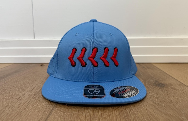 May include: A light blue baseball cap with a curved peak. The cap has six red, stylized chevrons embroidered across the front. The cap has a structured fit and a small, round button on top. The peak has a sticker.