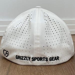 May include: White baseball cap with a perforated design. The cap has the text "GRIZZLY SPORTS GEAR" in black, and a logo. The hat is made of a lightweight material.