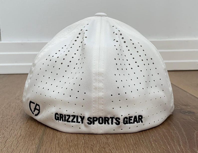 May include: White baseball cap with a perforated design. The cap has the text "GRIZZLY SPORTS GEAR" in black, and a logo. The hat is made of a lightweight material.