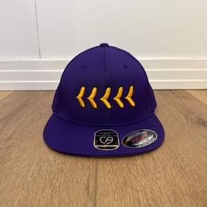 May include: A purple baseball cap with a curved peak. The cap has a series of yellow chevrons embroidered on the front. The cap has a structured fit and a fitted design.