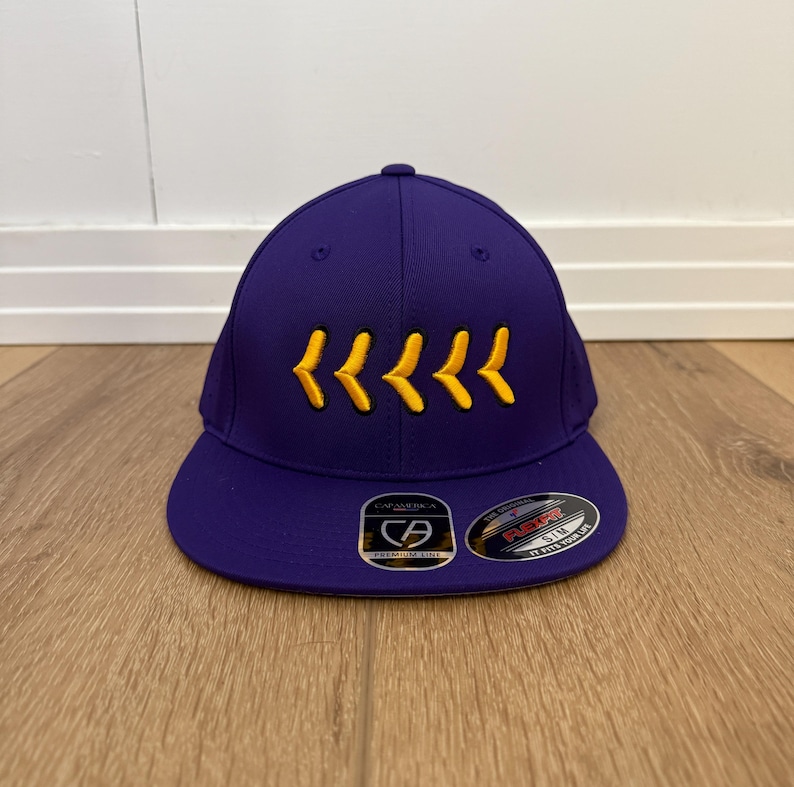 May include: A purple baseball cap with a curved peak. The cap has a series of yellow chevrons embroidered on the front. The cap has a structured fit and a fitted design.