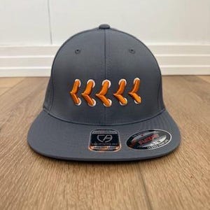 May include: A charcoal grey baseball cap featuring an embroidered design of five orange and white chevron-style laces across the front panels. The cap has a flat brim with two circular stickers attached, one showing a CA logo and the other a Flexfit label.