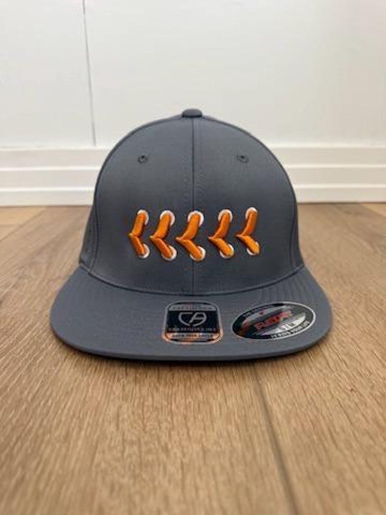 May include: A charcoal grey baseball cap featuring an embroidered design of five orange and white chevron-style laces across the front panels. The cap has a flat brim with two circular stickers attached, one showing a CA logo and the other a Flexfit label.