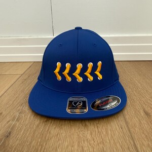 May include: A royal blue baseball cap with a curved peak. The cap has yellow embroidered chevrons across the front and a "Cap America" label. The hat is a Flexfit style.