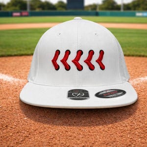 May include: White baseball cap with red stitching detail. The cap has a curved peak and a structured fit. The background shows a baseball field with green grass and a blue sky.