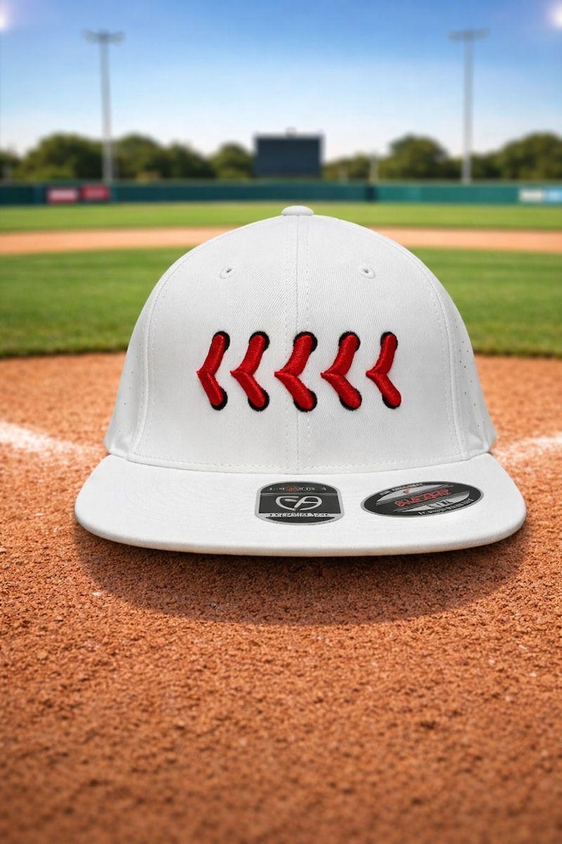May include: White baseball cap with red stitching detail. The cap has a curved peak and a structured fit. The background shows a baseball field with green grass and a blue sky.