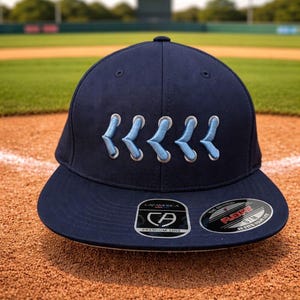 May include: Navy blue baseball cap with light blue embroidered detail. The cap has a curved peak and a structured fit. The cap features a "CAPAMERICA" logo and a "FLEXFIT" sticker.