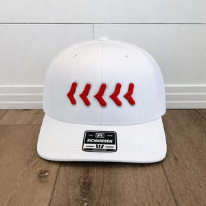 May include: A white baseball cap with a flat brim. The cap features five red chevrons embroidered across the front. The Richardson brand label is visible on the front of the brim. The cap is sitting on a wooden surface.