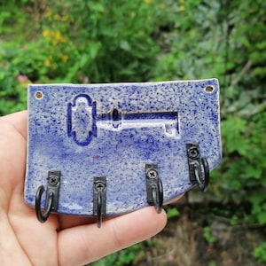 Może przedstawiać: Niebieski ceramiczny wieszak na klucze z motywem klucza i trzema czarnymi haczykami. Wieszak na klucze jest prostokątny z teksturowaną powierzchnią i dwoma otworami do zawieszenia. Tło to rozmyty widok zielonych liści.