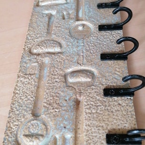 Può includere: Un portachiavi rettangolare con una superficie beige strutturata e disegni di chiavi in rilievo. È dotato di sei ganci in metallo nero per appendere le chiavi. Il portachiavi è lungo circa 25 cm e largo 13 cm.