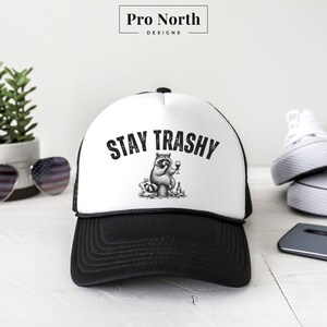 May include: Black and white trucker hat with a white front panel and a black mesh back. The front panel features a black and white illustration of a raccoon drinking wine and the text "STAY TRASHY".