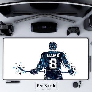 May include: A blue and black watercolor illustration of a hockey player wearing a jersey with the number 8 and the word "NAME" on the back. The player is holding a hockey stick and is facing away from the viewer. The background is white with blue and black splatters of paint.