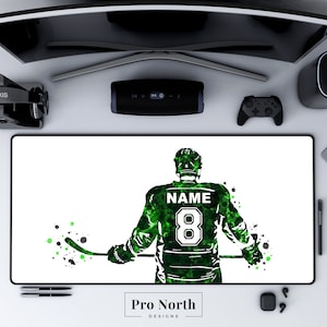 May include: Green and white hockey player silhouette with the text "NAME 8" on the back of the jersey. The player is holding a hockey stick and is surrounded by green and black paint splatters. The text "Pro North Designs" is at the bottom of the image.