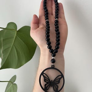 May include: Black beaded necklace with a butterfly pendant. The necklace features a series of round, glossy black beads and a circular pendant with a butterfly cutout design. The pendant is suspended from the beads.
