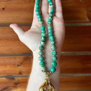 May include: A beaded necklace featuring large, round, green and tan jasper beads. The necklace has gold-colored accents and a gold-colored charm of boxing gloves.