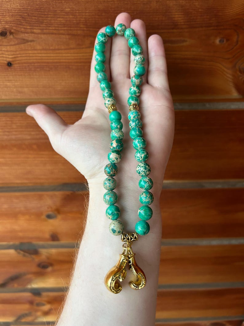 May include: A beaded necklace featuring large, round, green and tan jasper beads. The necklace has gold-colored accents and a gold-colored charm of boxing gloves.
