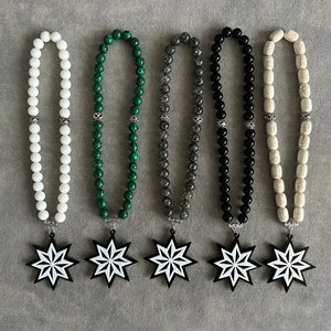 Puede incluir: Cinco rosarios o cuentas de oración con cuentas de diferentes colores y colgantes en forma de estrella. Las cuentas son blancas, verdes, grises, negras y blancas. Los colgantes en forma de estrella son negros con diseños blancos.