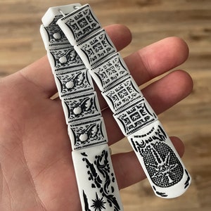 Puede incluir: Cuentas de oración blancas con diseños geométricos y florales negros. Las cuentas están enhebradas y tienen un borla al final.