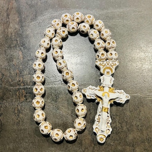 May include: A white rosary with gold detailing. The beads are decorated with a floral pattern, and the cross shows a detailed image of Jesus. This religious item is commonly used for prayer and meditation.