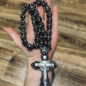 May include: Black prayer beads with a silver cross pendant. The beads are round and have a white star design. The cross has a silver crucifix and the inscription "IC XC NIKA" on the top.