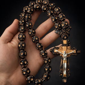 May include: A black rosary with gold detailing and a crucifix. The beads are round with a floral pattern. The crucifix has a gold cross and a silver Jesus figure.