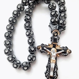 May include: A black and silver rosary with a detailed cross pendant. The beads are round and have a floral design. The cross features a silver figure of Jesus Christ.