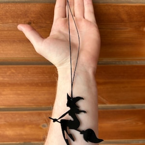 May include: Black witch silhouette pendant on a black cord. The witch is riding a broom. The pendant is hanging in front of a wooden background. The witch is wearing a pointed hat.