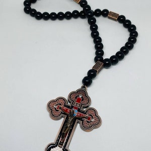 May include: Black beaded rosary with a large ornate copper cross featuring a depiction of Jesus Christ on the crucifix. The cross has red, blue, and white enamel accents.