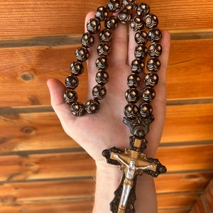 May include: A black wooden rosary with gold accents and a detailed wooden cross. The beads are round and have a floral pattern.