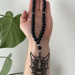May include: Black beaded necklace with a decorative black Oni mask pendant. The necklace features small, round beads and silver-colored accents. The Oni mask is a detailed, stylized design.