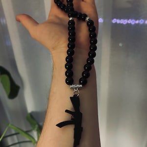 May include: Black beaded necklace with a silver-colored AK-47 pendant. The necklace features large, round, glossy black beads. The pendant is a detailed silhouette of an AK-47 assault rifle.