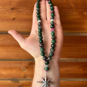 May include: A beaded necklace with green and pink stones, accented with silver-colored spacers. The necklace features a silver star pendant. The beads are a mix of green and pink, creating a unique color combination.