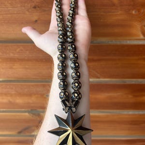 May include: A black and gold beaded necklace with a star pendant. The beads are black with gold accents, and the star pendant is black with gold detailing. The necklace is held up against a wooden background.