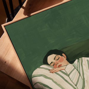 May include: An art print featuring a person sleeping, with closed eyes and rosy cheeks, resting on a striped pillow. The artwork is set against a dark green background and framed in a natural wood frame.