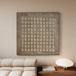 Peut inclure: Tableau abstrait encadré de bois carré. L'œuvre présente un fond texturé gris avec une grille de formes ovales blanc cassé. Il est accroché au-dessus d'un canapé clair et d'une table de chevet en bois.